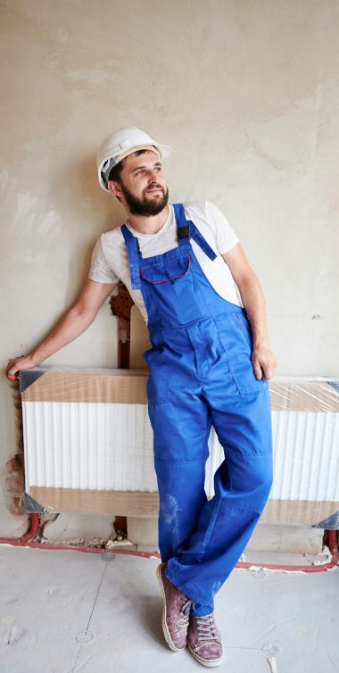 Young handsome plumber installing water radiator in unfinished apartment